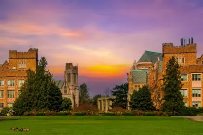 Gothic brick college buildings and evergreens behind a green lawn with geese at sunset