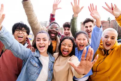 A diverse group of smiling young people outdoors, raising their hands and waving at the camera