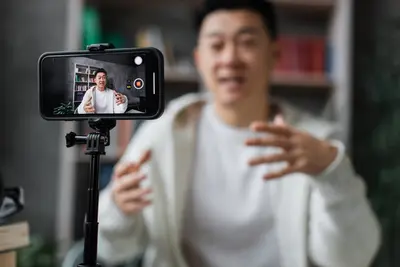 A smiling young man gestures as he records himself on a smartphone mounted on a tripod.