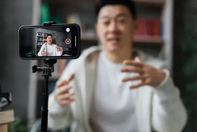 A smiling young man gestures as he records himself on a smartphone mounted on a tripod.