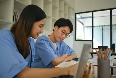 Two students in blue scrubs study together at a desk with a laptop, textbook and cup of pencils