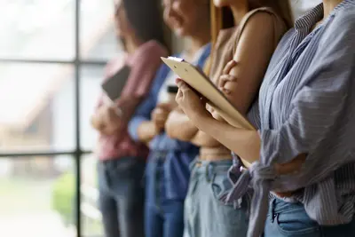 A line of students standing by a window, one holding a clipboard and pen while others wait.