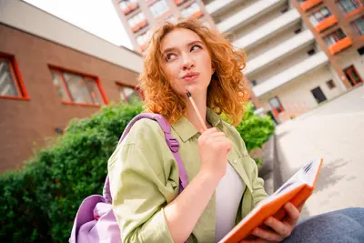 A student with curly red hair and a backpack holds a pencil to her chin and an open orange notebook, looking up thoughtfully outside a campus building.