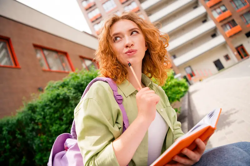 A student with curly red hair and a backpack holds a pencil to her chin and an open orange notebook, looking up thoughtfully outside a campus building.