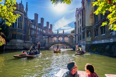 Punts carrying tourists along a sunlit river past historic stone college buildings and an ornate covered bridge
