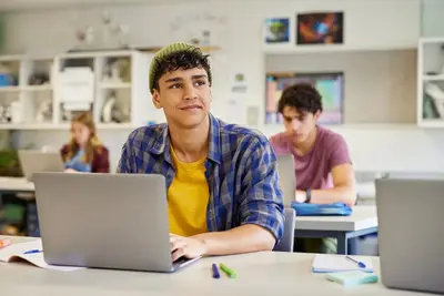 A high school student in a beanie and plaid shirt looks up thoughtfully from a laptop in a classroom.