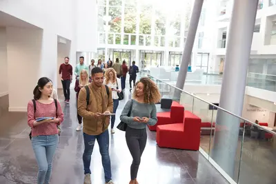 Students walking and chatting in a bright, modern university lobby with red sofas and tall windows