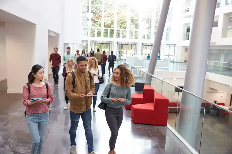 Students walking and chatting in a bright, modern university lobby with red sofas and tall windows