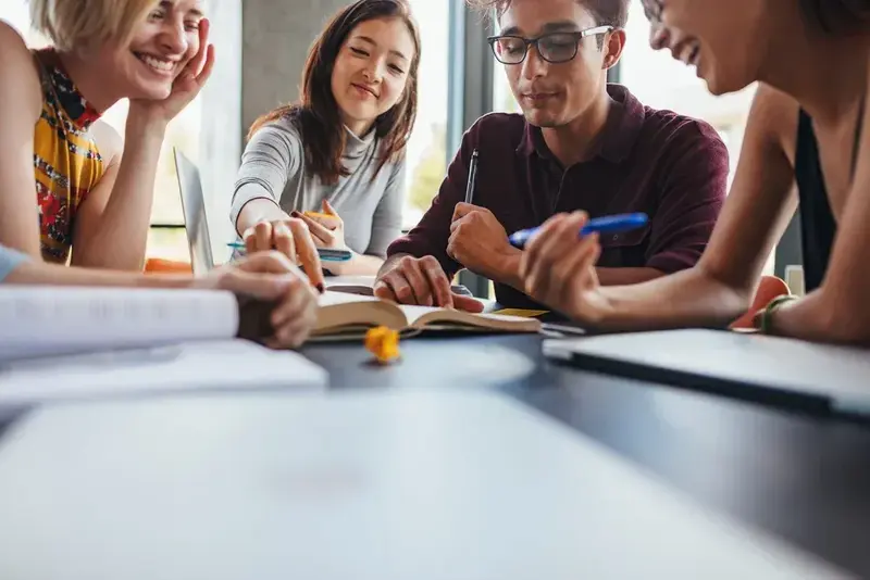Four smiling students leaning over a table with an open book, notebooks and pens