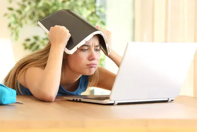 A frustrated student holds a notebook over her head while staring at a laptop screen at a desk.