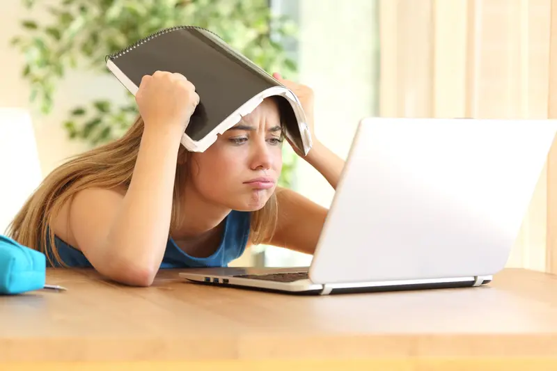 A frustrated student holds a notebook over her head while staring at a laptop screen at a desk.