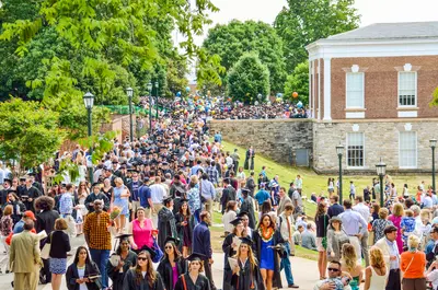 Large crowd of graduates in caps and gowns and families filling a tree-lined campus walkway on graduation day