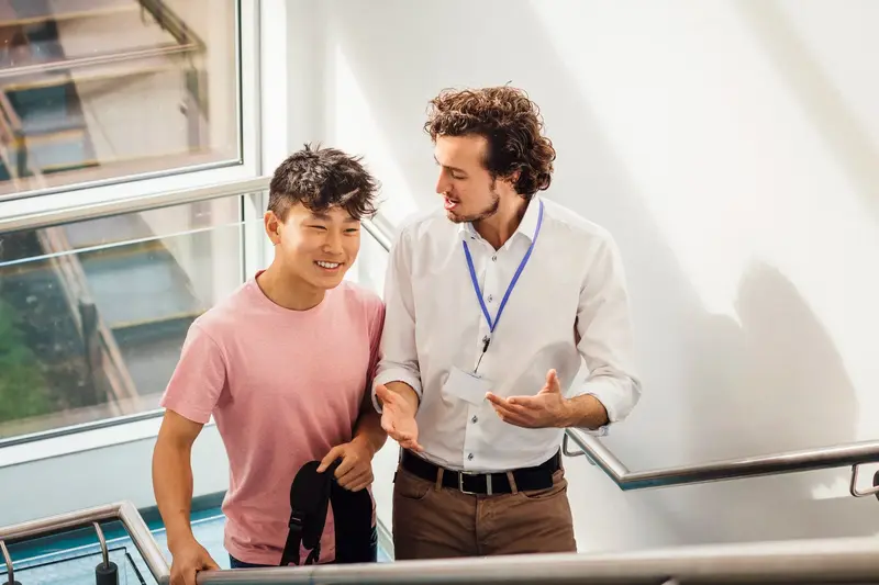 A staff member wearing a lanyard talks with a smiling student on a stairway by a window