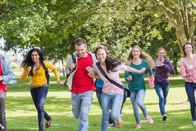A group of smiling teenagers with backpacks running across a sunny park lawn beneath leafy trees