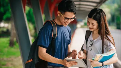 Two students with backpacks looking at a tablet together outdoors, one holding books
