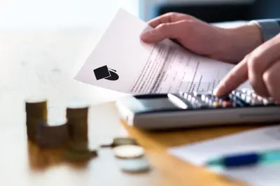 A person holding a document marked with a graduation cap icon while using a calculator beside stacks of coins