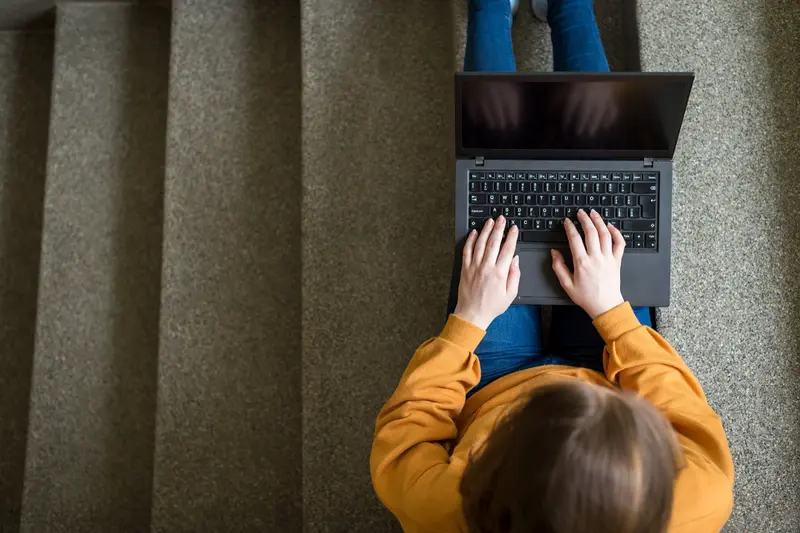 Overhead view of a student in an orange sweater typing on a laptop while sitting on carpeted stairs