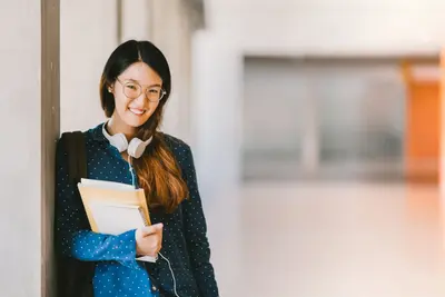 Smiling student in glasses with headphones around her neck holding notebooks in a school hallway