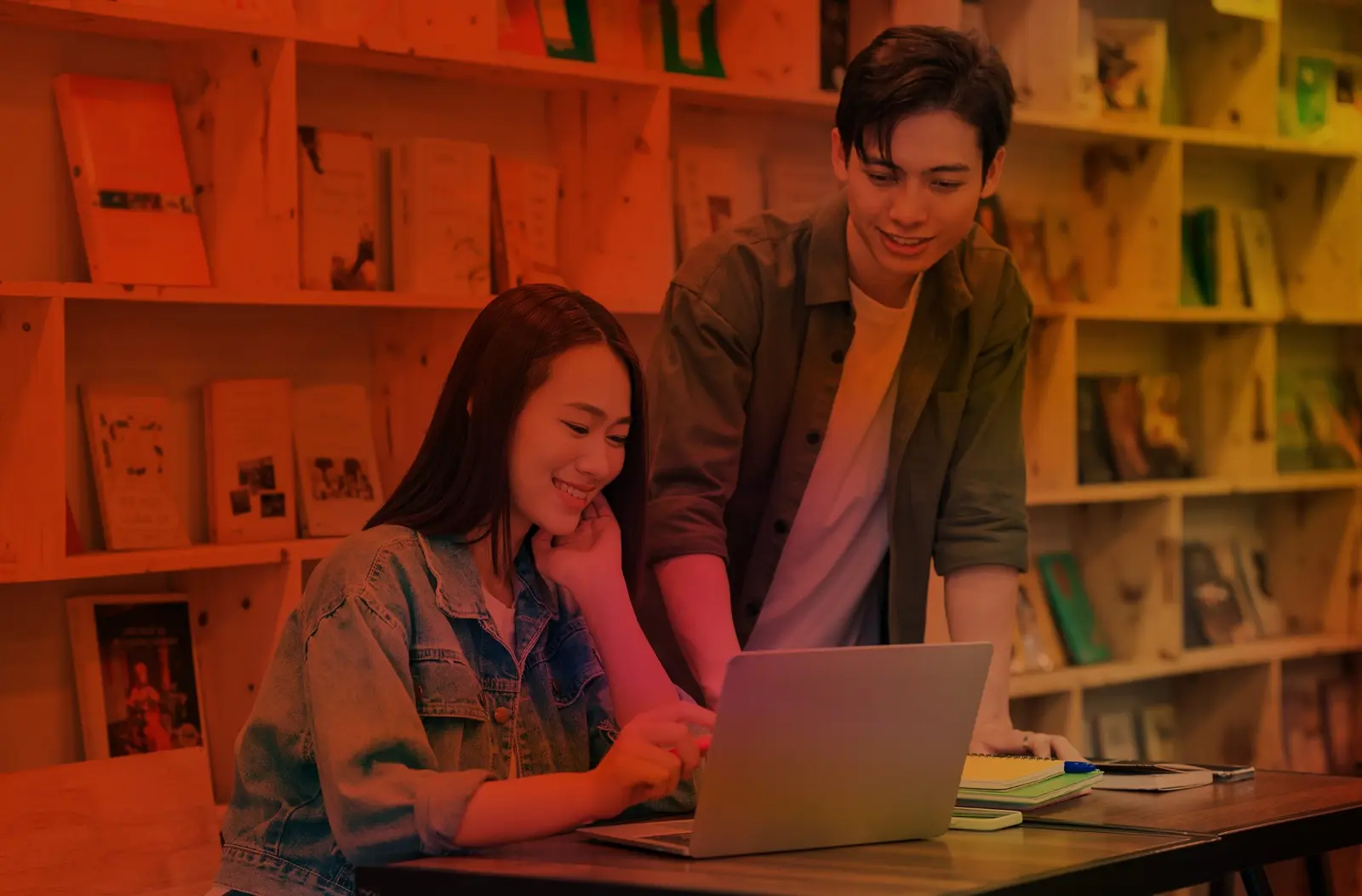 Two smiling students looking at a laptop together at a table in front of bookshelves