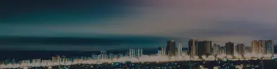 A coastal city skyline of high-rise buildings lit up at dusk under a hazy teal and pink sky