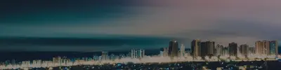 A coastal city skyline of high-rise buildings lit up at dusk under a hazy teal and pink sky