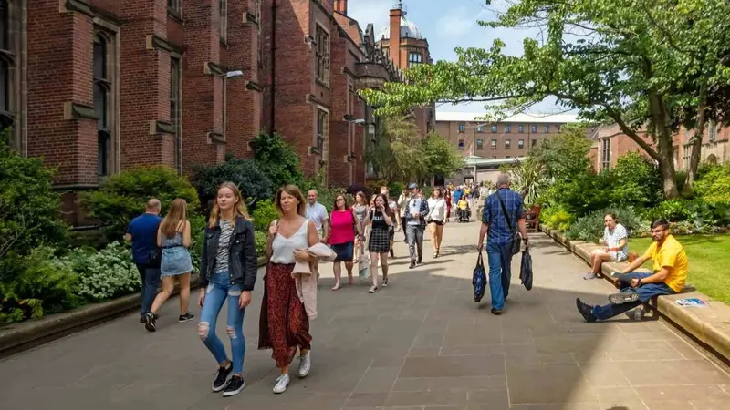 Students walking and sitting along a tree-lined campus path beside a red brick university building.