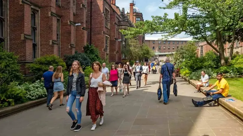 Students walking and sitting along a tree-lined campus path beside a red brick university building.