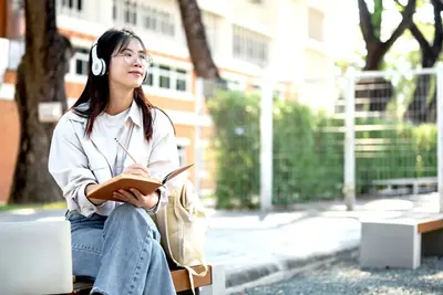 A student wearing headphones sits on an outdoor bench with a notebook and pencil, looking up thoughtfully.