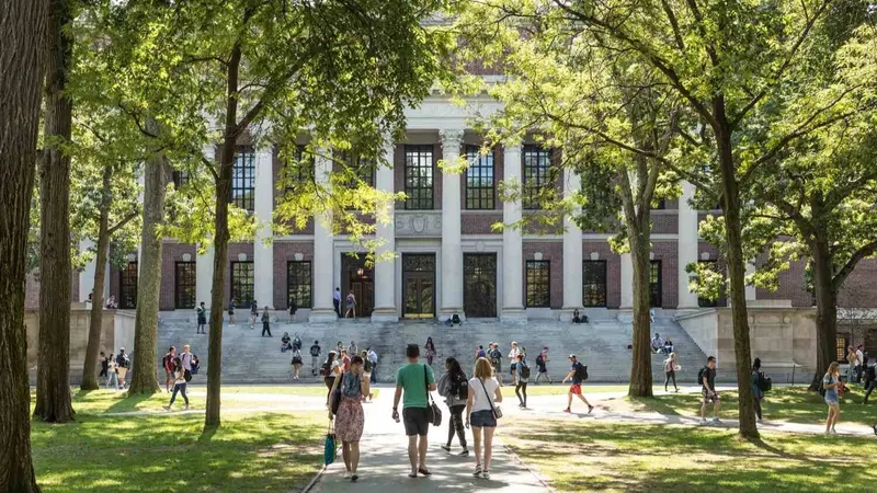 Students walking across a sunny tree-lined campus lawn toward a large columned brick library building.