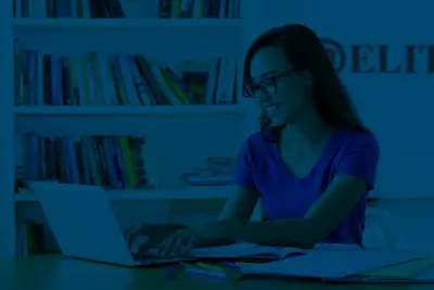 A smiling student in glasses types on a laptop at a desk with notebooks, in front of bookshelves