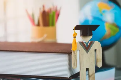 A small wooden figure wearing a graduation cap stands beside stacked books, colored pencils, and a globe.