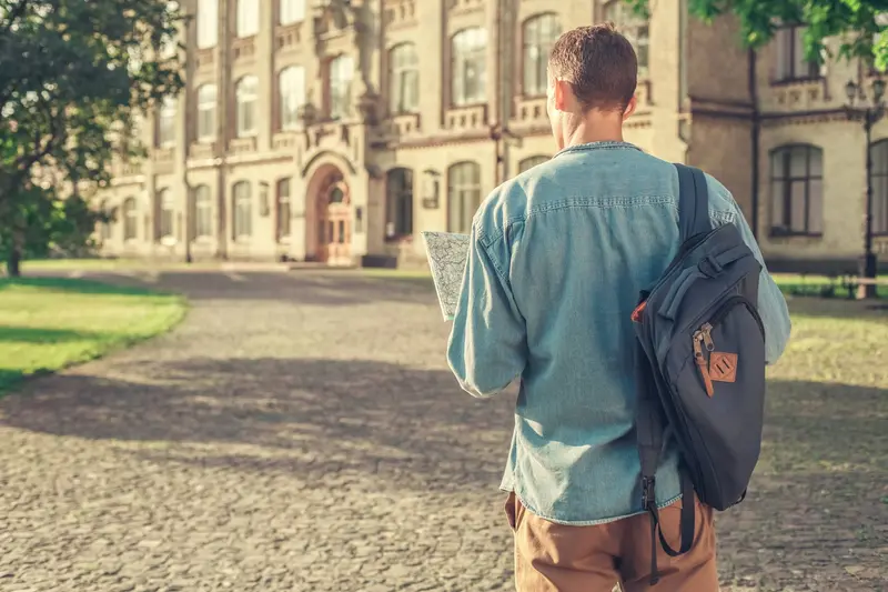 A student with a backpack holds a map while walking toward a large stone campus building.