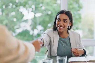 A smiling young woman in a blazer shakes hands with someone across a table by a sunny window