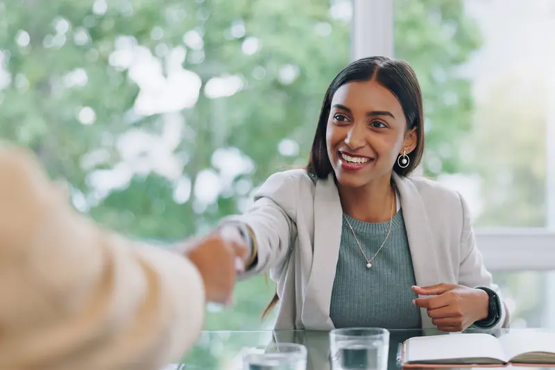 A smiling young woman in a blazer shakes hands with someone across a table by a sunny window