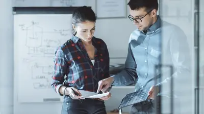 Two students reviewing papers and a tablet together in front of a whiteboard covered in diagrams