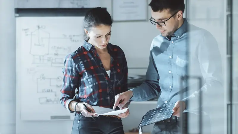 Two students reviewing papers and a tablet together in front of a whiteboard covered in diagrams