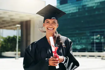 A smiling graduate in cap and gown holds a diploma tied with a red ribbon outdoors near city buildings.