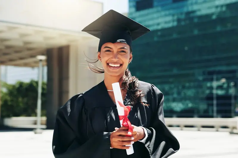 A smiling graduate in cap and gown holds a diploma tied with a red ribbon outdoors near city buildings.