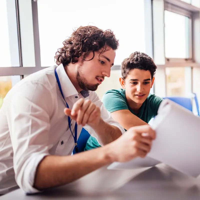A teacher wearing a lanyard points at a sheet of paper while a teenage student reads along beside him