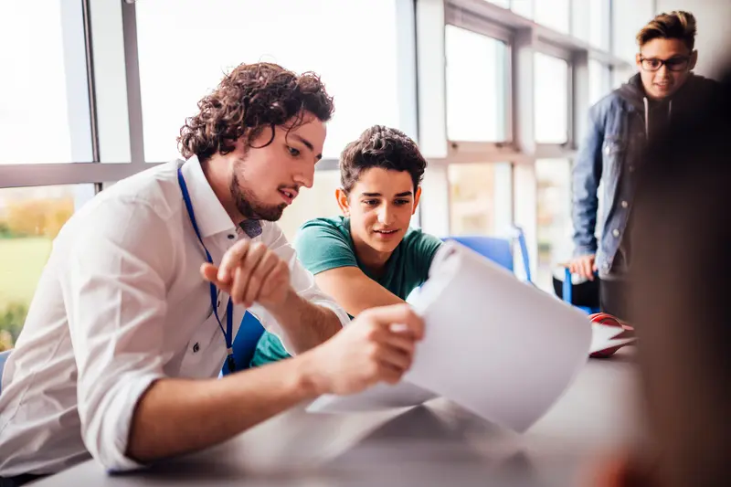 A teacher and a student lean over a desk reviewing papers together while another student watches nearby