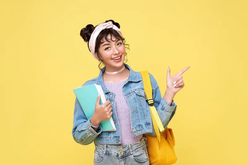 Smiling student with a yellow backpack holding notebooks and pointing to the side against a yellow background