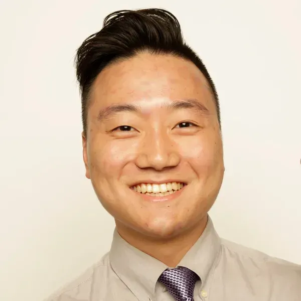 Smiling young man in a tan dress shirt and patterned purple tie against a plain background