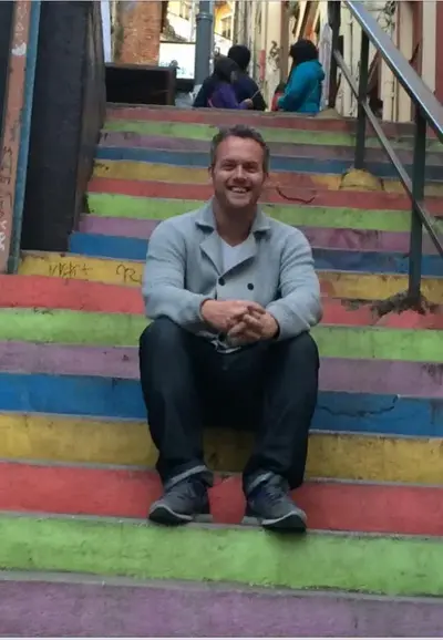 Smiling man in a gray cardigan sitting on a staircase painted in rainbow stripes.