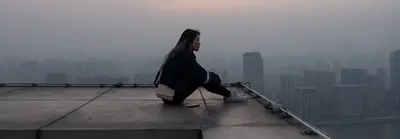 해질 무렵 옥상 가장자리에 홀로 앉아 안개 낀 도시 스카이라인을 바라보는 젊은 여성의 모습입니다.