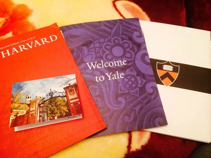 Three college acceptance packets fanned out: a red Harvard folder, a purple "Welcome to Yale" booklet, and a white Princeton crest envelope.