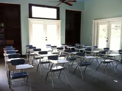 Empty classroom with rows of blue tablet-arm desks facing bright French doors, bookshelves along the walls