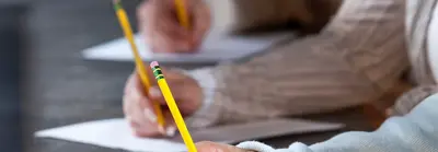 Close-up of two students holding yellow pencils as they write on papers at a desk