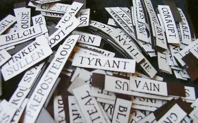 TYRANT, VAIN, CORRUPT, BELCH, SULLEN, PREPOSTEROUS 등의 단어가 적힌 자석 단어 조각들이 흩어져 쌓여 있는 모습