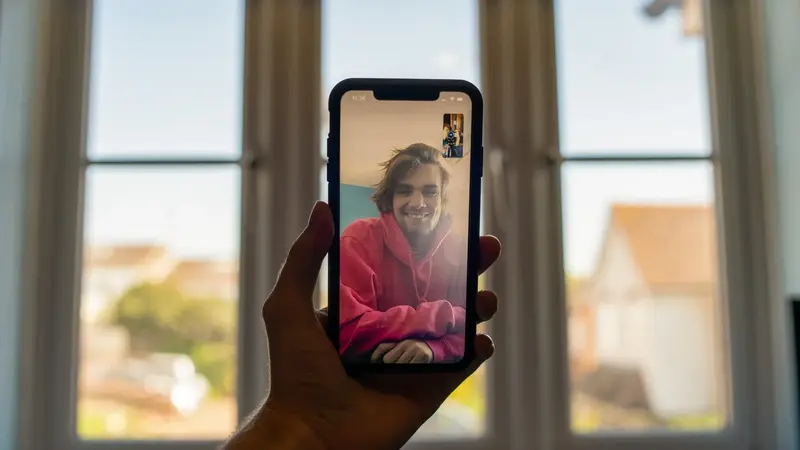 A hand holds a smartphone showing a video call with a smiling person in a pink hoodie, in front of a bright window.