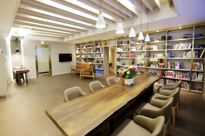 Study lounge with a long wooden table, cushioned chairs, pendant lights, and walls of filled bookshelves.